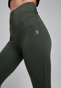 Hoge taille sportlegging in donker groen, gemaakt van een gladde stof. Heeft stikseldetails en een klein wit logo aan de zijkant.