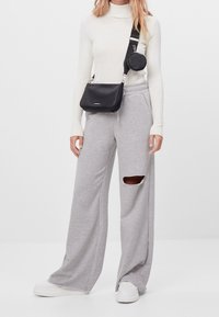 Kobieta w białym, prążkowanym golfie, jasnoszarych szerokich spodniach dresowych z wycięciem na kolano, białych sneakersach i czarnej torbie na ramię z okrągłym woreczkiem.