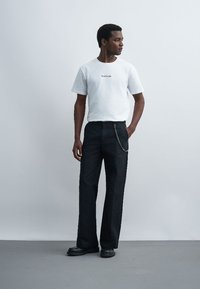 Homme debout avec une main dans la poche, portant un t-shirt blanc à manches courtes et un pantalon noir à jambes larges avec un accessoire chaîne.