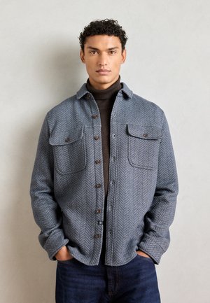 Gray herringbone shirt with two chest pockets, button front, and rounded collar, layered over a dark turtleneck sweater. Casual fit.