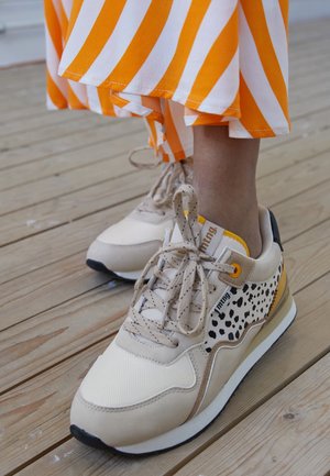 Fødder iført beige sneakers med sorte pletter og gule accenter, orange og hvide stribede nederdel, stående på træplankegulv.