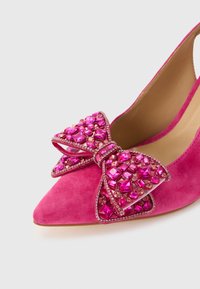 Roosa seemisnahast teravate ninadega kõrged kontsad, millel on ees suur dekoratiivne lips, mis on kaunistatud erkroosade kivikestega.