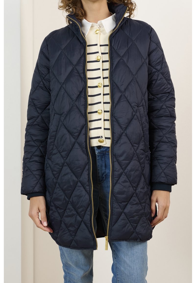 Chaqueta acolchada de puffer larga en color azul marino con cuello alto, cierre de cremallera dorado y puños acanalados. Lleva puesta sobre una blusa a rayas y jeans de mezclilla.