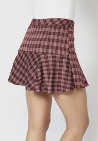 Mini-jupe évasée à carreaux bordeaux et blancs portée sur les hanches basses, montrant partiellement les jambes, sur un fond uni.