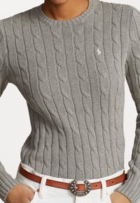 Personne portant un pull en maille torsadée gris rentré dans un pantalon blanc avec une ceinture en cuir marron ornée d'une boucle en argent décorative.