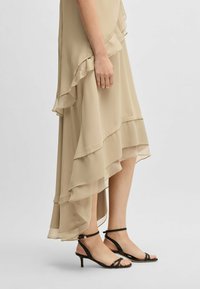 Beige chiffon-mekko, jossa on kerrokselliset räpylät helmassa, yhdistetty mustiin kapeisiin korkokenkiin, joissa on ohut nilkkaremmi ja matala korko.