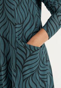 Teal jurk met zwart loofpatroon, voorzien van een zijzak en aansluitende mouwen. Gemaakt van zachte stof met een gladde textuur.