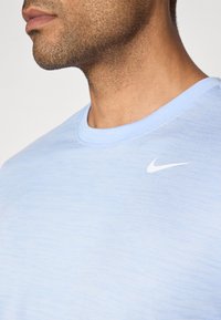 Svetlo modra Nike majica s gladko teksturo, okroglo ovratnico in belim logotipom na zgornjem levem prsnem delu. Preprost dizajn, brez vzorcev ali dodatkov.