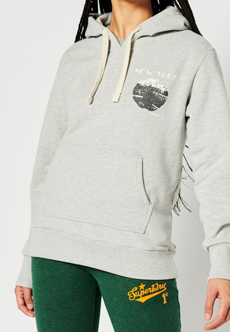 Personne portant un sweat à capuche gris clair avec motif New York et un pantalon de survêtement SuperDry vert foncé, debout devant un fond blanc.