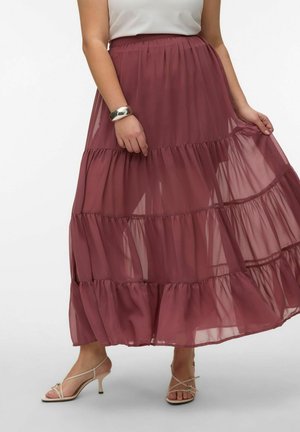 Žena má na sebe priesvitnú viacvrstvovú maxi sukňu v fialkovom odtieni, biely tielkový top, strieborný náramok a béžové sandále na podpätku s remienkami, drží okraj sukne.