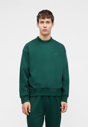 Sweatshirt verde com gola redonda em canelado, ombros caídos e mangas com punhos. Apresenta um logo subtil no peito. Textura de tecido suave.