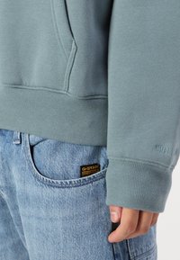 Felpa azzurra con orlo e polsini a coste, dotata di tasca a marsupio. Indossata con jeans in denim blu sbiaditi e un'etichetta visibile.
