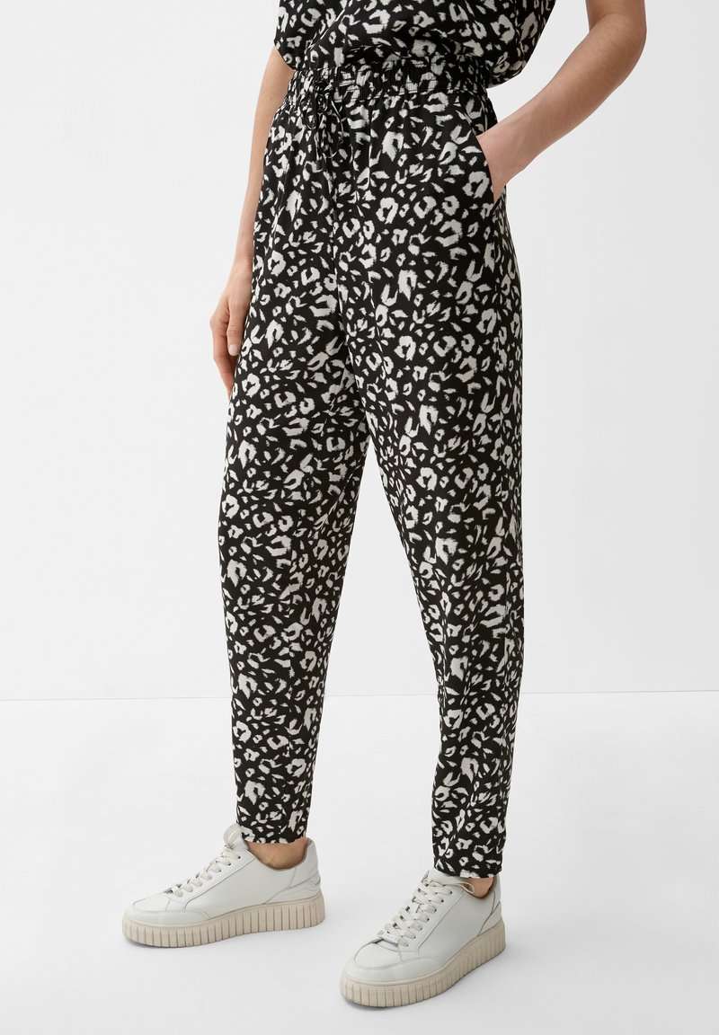 Personne portant un pantalon à imprimé léopard noir et blanc avec une taille élastique et des baskets blanches, se tenant avec une main dans la poche.