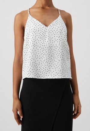 Frau trägt ein weißes ärmelloses V-Ausschnitt-Oberteil mit schwarzen Punkten und einen hoch taillierten schwarzen Rock, vor einem einfarbigen Hintergrund.