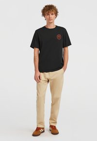 Black cotton t-shirt with short sleeves and chest graphic. Paired with beige chinos and brown sneakers featuring red laces and white stripes.