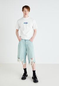 Tricou alb cu mânecă scurtă și grafică colorată, asociat cu șorturi din denim de lungime medie, de culoare albastru deschis, cu margini desfăcute și detalii rupte.