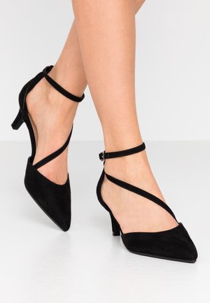 Czarne zamszowe buty na wysokim obcasie z noskiem w szpic i krzyżującymi się paskami wokół kostki. Obcas ma około 7,5 cm wysokości. Gładka faktura na całej powierzchni.