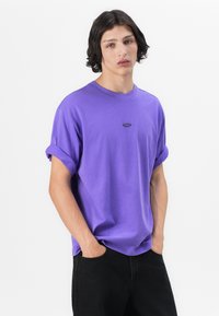 Purple short-sleeve t-shirt with rolled cuffs and a small brand logo on the chest, made of soft cotton material.
