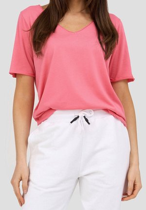 Femme portant un t-shirt ample rose à manches courtes et col en V, rentré dans un pantalon blanc à cordon, sur un fond uni.