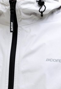 Veste blanche avec une fermeture éclair noire et un curseur étiqueté "JJcore", dotée d'une capuche avec un cordon noir réglable et un bouton-poussoir en plastique.