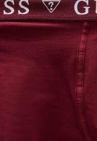 Κοντινό πλάνο σε βυσσινί ύφασμα με λεπτομέρεια ραφής και διπλωμένη ζώνη μέσης που δείχνει μερικό λευκό κείμενο, περιλαμβάνοντας τα γράμματα "SS" και ένα τρίγωνο με ερωτηματικό.