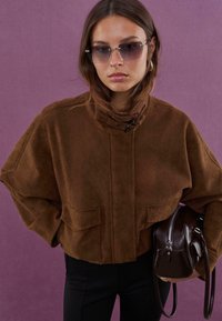 Veste courte en suède marron avec col haut et deux poches avant ; associée à un pantalon noir et un petit sac à main sombre. Fond violet.