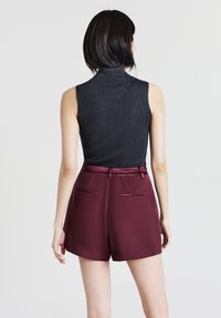 Donna di spalle che indossa un dolcevita nero a coste senza maniche e pantaloni corti bordeaux a vita alta con tasche a filetto sul retro, su uno sfondo semplice.