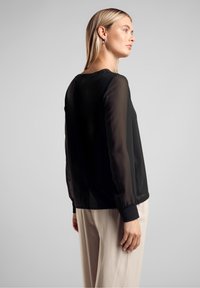 Blusa nera con maniche lunghe trasparenti, scollatura rotonda e polsini elastici. Abbinata a pantaloni chiari, mostrata da dietro.