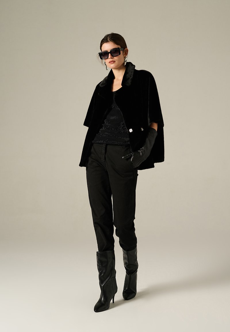 Black faux fur cape jacket with a collar, worn over a black sequin top, paired with black pants and knee-high heeled boots. Black sunglasses worn.