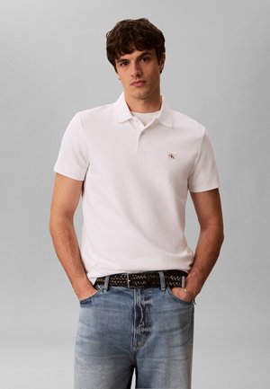 Fiatal férfi fehér CK logós galléros pólót, világoskék farmert és fekete szőtt övet visel, zsebében tartja a kezét, szürke háttér előtt áll.