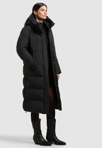 Manteau rembourré noir long avec capuche, présentant une coupe ample, une fermeture éclair à l'avant et des poches latérales. Porté avec des bottes noires jusqu'aux genoux.
