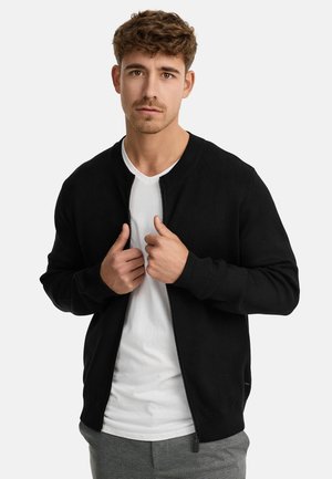 Suéter negro con cremallera hecho de una tela suave, con cuello redondo y puños de canalé, superpuesto a una camiseta blanca de cuello en V.