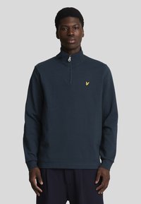 Pullover a quattro zip di colore blu navy realizzato in tessuto morbido, con maniche lunghe e un piccolo logo di un'aquila gialla sul petto.