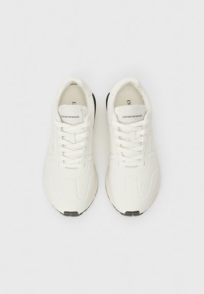 Valged tennisemed tekstuuriga sünteetilisest ülemisest, lamedad paelad, ümar varvas ja must kummist tald. Brändi logo keele peal.