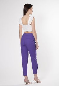 Witte crop top met kantdetails en ruches op de schouderbanden, gecombineerd met paarse tapered broek met zijzakken en knoopaccenten.