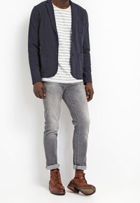 Hombre con blazer azul marino, camisa blanca con rayas grises, jeans grises remangados y botas de cuero marrón, de pie frente a un fondo blanco.