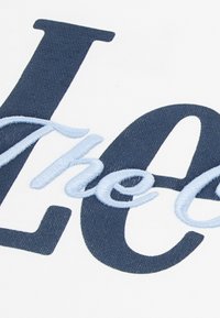 Λευκό ύφασμα εμφανίζει μεγάλα, ναυτικά μπλε γράμματα με ελαφρώς μπλε, κεντημένες λεπτομέρειες. Το κείμενο συνδυάζει στιλ έντονα και καλλιγραφικά.