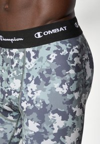 Feszes leggings fekete derékrésszel, amelyen a "Champion" és a "COMBAT" felirat található. Az anyag szürke és világoskék álcázó mintával rendelkezik.