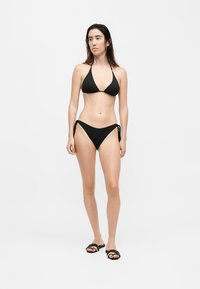Jeune femme aux longs cheveux foncés portant un bikini noir et des sandales slides noires, debout devant un fond uni gris clair.
