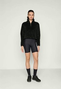 Veste zippée en polaire noire, col montant, associée à un short de cyclisme à motifs sombres. Des chaussettes et des chaussures de sport noires complètent la tenue.