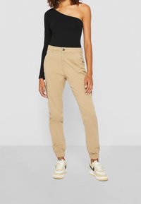 Mustassa yhden olkapään bodysuitissa, johon on yhdistetty beigeet cargo-housut, joissa on elastiset resorit ja sivutaskut. Asustuksen täydentävät valkoiset Nike-tennarit.