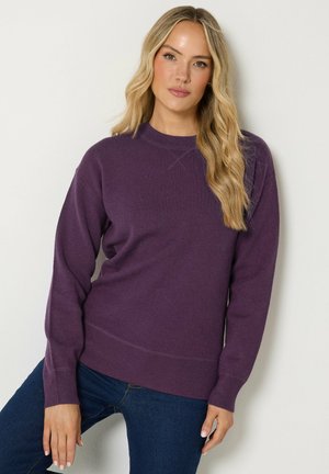 Femme aux longs cheveux blonds portant un pull violet à manches longues et un jean bleu sur un fond clair uni.
