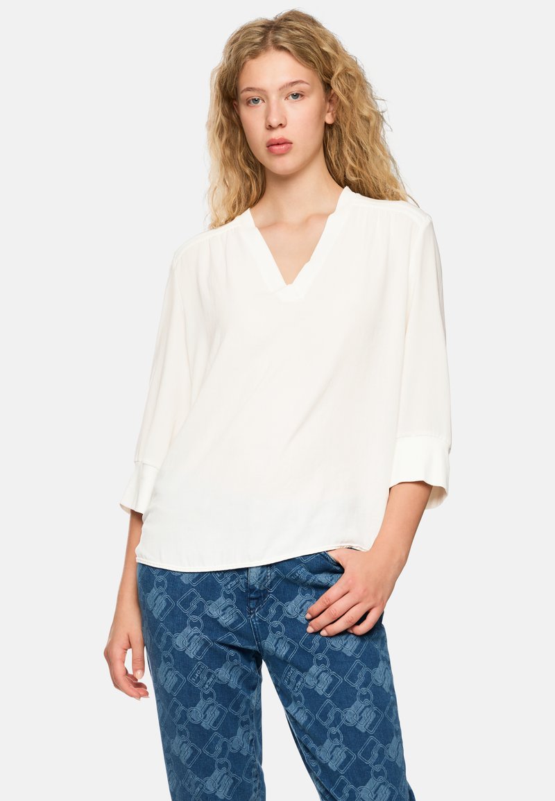 White, loose-fitting blouse with a V-neck and three-quarter sleeves. Soft fabric, smooth texture. Paired with blue patterned jeans.