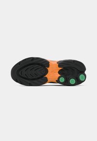 Sort og orange sneaker sål med tekstureret slidbane og tre grønne cirkulære elementer nær hælen på en hvid baggrund.