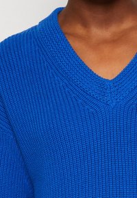 Pull tricoté bleu vif avec un col en V, présentant un tissu texturé et des détails côtelés le long de l'encolure.