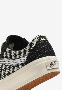 Sneaker in bianco e nero con motivo a tessuto intrecciato, collo imbottito, accenti bianchi e suola beige con un'etichetta grigia sul lato.
