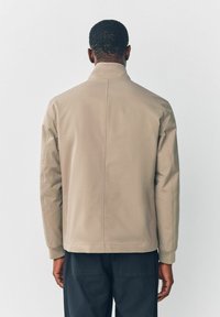 Beige jakke med høj krave, lange ærmer og afslappet pasform. Glat tekstur, minimale sømme og ribbede manchetter.