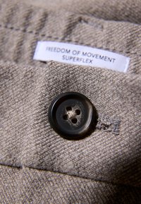 Beige stof met een gestructureerde weving, voorzien van een donkere knop met vier gaten. Het label leest "FREEDOM OF MOVEMENT SUPERFLEX."