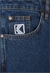 Jeans iz jeansa temno modre barve. Imajo majhno zanko z belim vezenim logotipom, zlatim okovjem in pomarančnimi šivi.