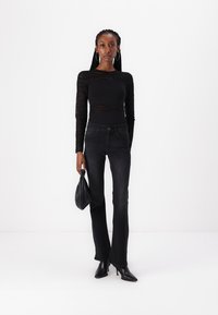 Schwarzes langärmliges Oberteil aus transparenter Stoff, kombiniert mit hochgeschnittenen schwarzen Jeans mit dezenten Verzierungen und schwarzen Absatzschuhen. Trägt eine schwarze Tasche.